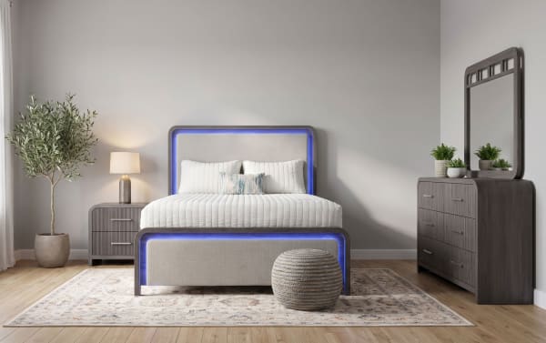 Modern bedroom with a beige upholstered bed featuring blue LED frame lighting, gray nightstand with lamp, dark wood dresser with mirror, and indoor plant on light wood flooring.